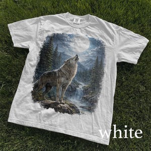 May include: White t-shirt featuring a detailed graphic of a howling wolf in a forest scene under a full moon. The design includes a waterfall and evergreen trees, creating a nature-themed image. The word "white" is printed at the bottom.