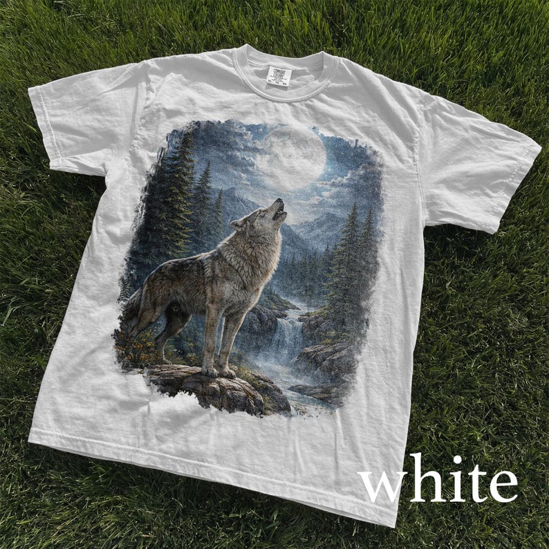 May include: White t-shirt featuring a detailed graphic of a howling wolf in a forest scene under a full moon. The design includes a waterfall and evergreen trees, creating a nature-themed image. The word "white" is printed at the bottom.