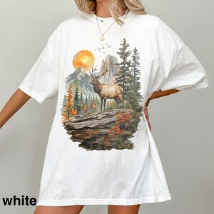 K&ouml;nnte beinhalten: Wei&szlig;es T-Shirt mit einem Naturmotiv, das einen Elch, Berge und B&auml;ume zeigt. Die Grafik enth&auml;lt eine untergehende Sonne und mehrere kleinere Hirsche. Das Wort "white" ist unten auf dem Shirt aufgedruckt.
