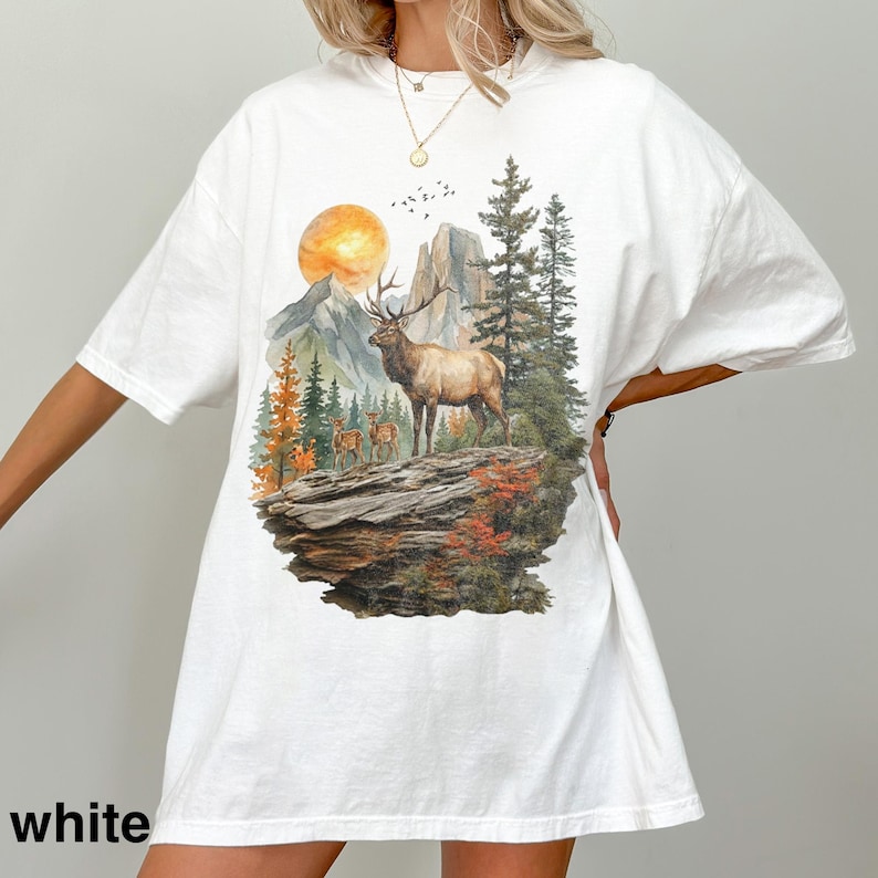K&ouml;nnte beinhalten: Wei&szlig;es T-Shirt mit einem Naturmotiv, das einen Elch, Berge und B&auml;ume zeigt. Die Grafik enth&auml;lt eine untergehende Sonne und mehrere kleinere Hirsche. Das Wort "white" ist unten auf dem Shirt aufgedruckt.
