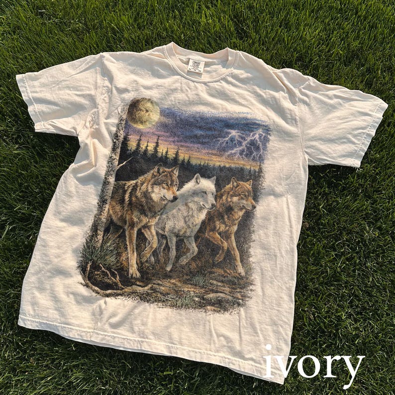 May include: Ivory-colored t-shirt featuring a detailed graphic of three wolves in a forest setting under a stormy sky with lightning and a full moon. The wolves are depicted in shades of brown and white.