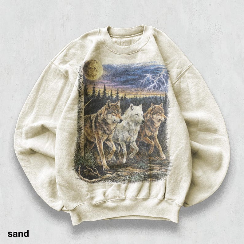 May include: Sand-colored sweatshirt featuring a graphic of three wolves in a forest scene under a stormy sky with lightning and a full moon. The wolves are depicted in shades of brown and white.