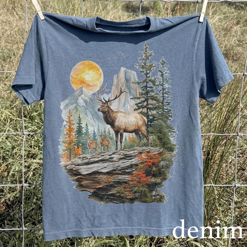 K&ouml;nnte beinhalten: Blaues Kurzarm-T-Shirt mit einer Naturszene mit einem gro&szlig;en Elch, kleineren Hirschen, Bergen und B&auml;umen unter einer gelben Sonne. Das Wort "denim" ist unten aufgedruckt.