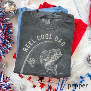 May include: A stack of folded t-shirts in red, white, and light blue. The top shirt is pepper gray and features the text "REEL COOL DAD" with a graphic of a fish and fishing rod. The shirts are on a white, fluffy surface.