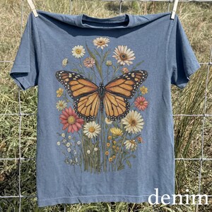 Puede incluir: Una camiseta azul con un dise&ntilde;o de mariposa monarca, con alas naranjas y negras, sobre un fondo de flores vibrantes. La camiseta tiene cuello redondo y mangas cortas. La palabra "denim" est&aacute; impresa en la parte inferior.