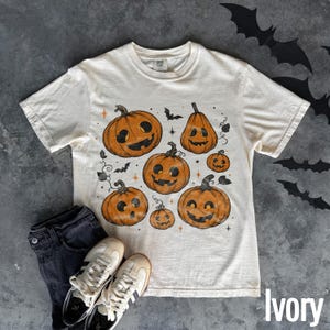 May include: An ivory short-sleeve t-shirt featuring a graphic print of six orange jack-o'-lanterns with various facial expressions, surrounded by small black bats and orange star accents. The shirt has a crew neckline and a standard fit.