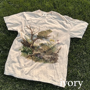 May include: Ivory-colored t-shirt featuring a detailed graphic of two green frogs perched on a log with surrounding foliage. The design includes a tree branch, ferns, and wildflowers, creating a nature-themed aesthetic.