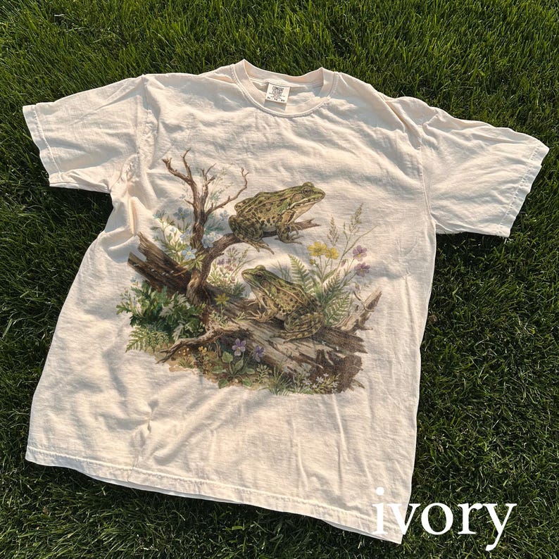 May include: Ivory-colored t-shirt featuring a detailed graphic of two green frogs perched on a log with surrounding foliage. The design includes a tree branch, ferns, and wildflowers, creating a nature-themed aesthetic.