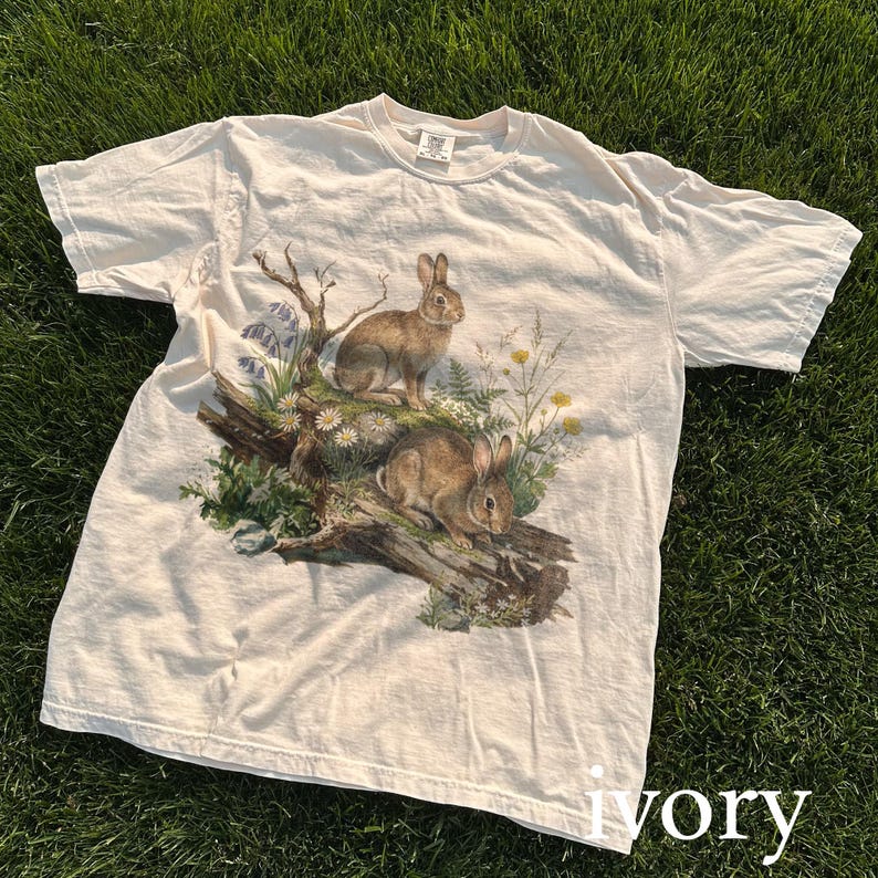 K&ouml;nnte beinhalten: Elfenbeinfarbenes T-Shirt mit einer detaillierten Illustration von zwei braunen Kaninchen inmitten von Wildblumen und Bl&auml;ttern auf einem gefallenen Baumstamm. Das Design umfasst blaue und gelbe florale Akzente und schafft eine von der Natur inspirierte &Auml;sthetik.