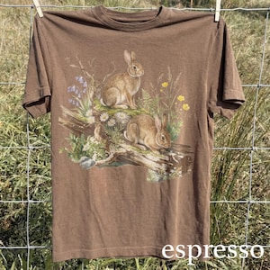 K&ouml;nnte beinhalten: Braunes T-Shirt mit einer detaillierten Illustration von zwei Kaninchen inmitten von Wildblumen und Bl&auml;ttern. Die Grafik enth&auml;lt blaue, gelbe und wei&szlig;e Blumen, mit dem Wort "espresso" unten aufgedruckt. Das T-Shirt h&auml;ngt an einer W&auml;scheleine.