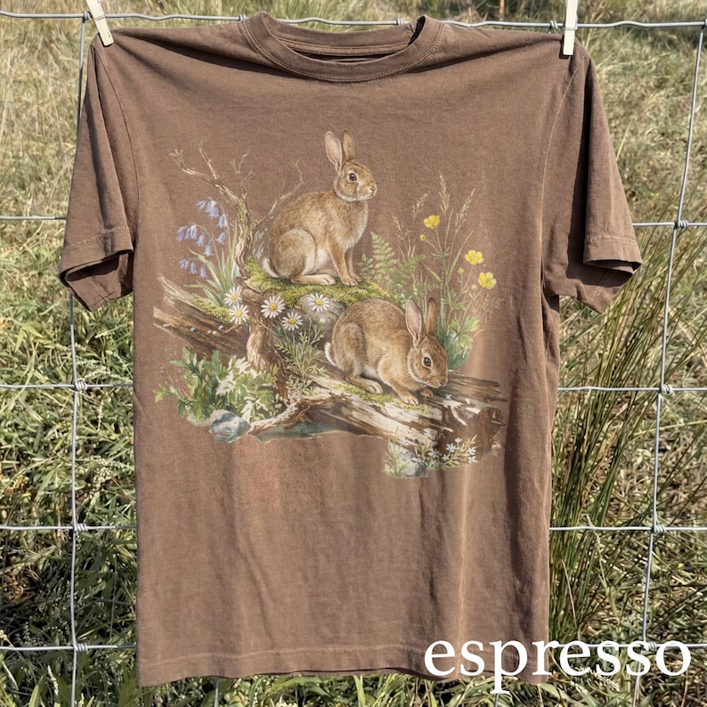 K&ouml;nnte beinhalten: Braunes T-Shirt mit einer detaillierten Illustration von zwei Kaninchen inmitten von Wildblumen und Bl&auml;ttern. Die Grafik enth&auml;lt blaue, gelbe und wei&szlig;e Blumen, mit dem Wort "espresso" unten aufgedruckt. Das T-Shirt h&auml;ngt an einer W&auml;scheleine.