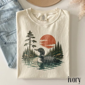 May include: An ivory short-sleeve t-shirt featuring a graphic print of a loon swimming on a lake, surrounded by a forest of pine trees and a large orange sun. The inner neck label displays the text Comfort Colors and the size M.
