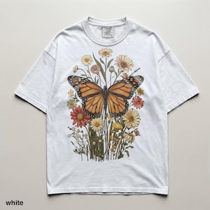 Puede incluir: Camiseta blanca con una ilustraci&oacute;n en acuarela de una mariposa monarca con alas naranjas y negras, rodeada de flores coloridas. La camiseta tiene cuello redondo y mangas cortas. El texto "TIMELESS COLORS" est&aacute; impreso en la parte superior izquierda.