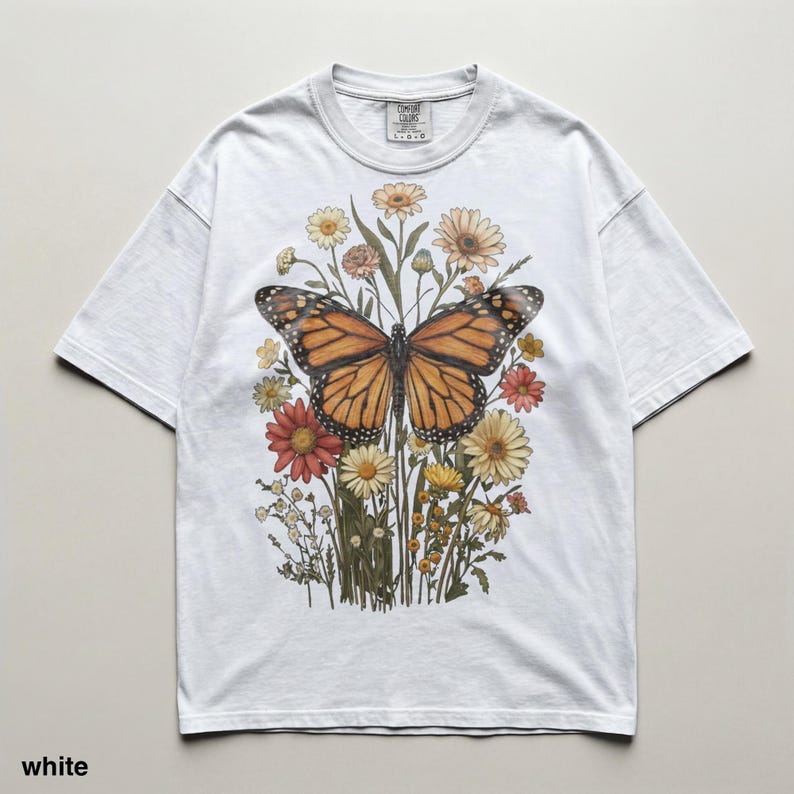 Puede incluir: Camiseta blanca con una ilustraci&oacute;n en acuarela de una mariposa monarca con alas naranjas y negras, rodeada de flores coloridas. La camiseta tiene cuello redondo y mangas cortas. El texto "TIMELESS COLORS" est&aacute; impreso en la parte superior izquierda.