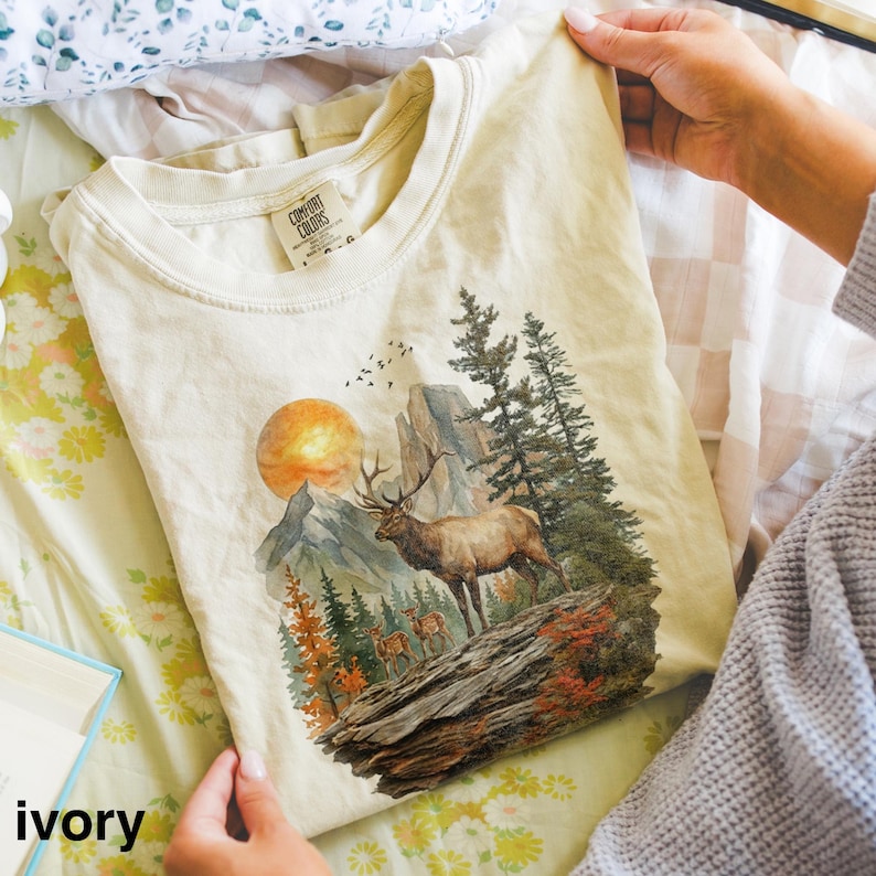 K&ouml;nnte beinhalten: Elfenbeinfarbenes T-Shirt mit einer Berglandschaft, einem Elch, B&auml;umen und einer untergehenden Sonne. Das Shirt hat einen Rundhalsausschnitt und kurze &Auml;rmel. Das Wort "ivory" ist unten aufgedruckt.
