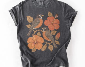 Puede incluir: Una camiseta de manga corta gris carbón con un estampado gráfico de dos pájaros marrones y naranjas posados en ramas, rodeados de grandes flores de hibisco naranjas y hojas. La prenda tiene cuello redondo y una textura de tela desgastada.