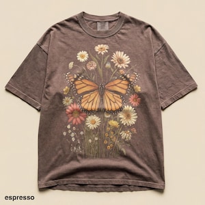Puede incluir: Camiseta color espresso con una gran mariposa de alas naranjas y negras, rodeada de flores coloridas. El dise&ntilde;o est&aacute; centrado en la parte delantera de la camiseta. La camiseta tiene mangas cortas y cuello redondo.