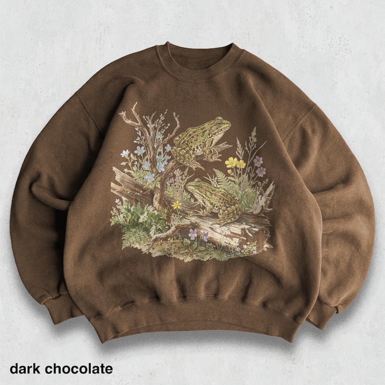 May include: Dark chocolate-colored sweatshirt featuring a detailed illustration of two frogs perched on a log with plants and flowers. The text "dark chocolate" is printed at the bottom. The sweatshirt has a crew neck and long sleeves.