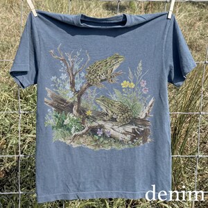May include: Blue denim t-shirt featuring a detailed illustration of two frogs perched on a log with wildflowers. The frogs are green and brown, and the shirt has a classic crew neck design. The word "denim" is printed at the bottom.