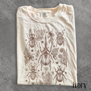May include: Ivory-coloured t-shirt with a brown illustration of beetles, leaves, acorns, and stars. The shirt has a crew neck and is made of a soft material. The word "ivory" is printed at the bottom right. The shirt is folded.