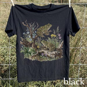 May include: Black t-shirt featuring a detailed illustration of two frogs perched on a log surrounded by plants and flowers. The design includes a variety of colors, including green, blue, and yellow. The word "black" is printed at the bottom of the shirt.