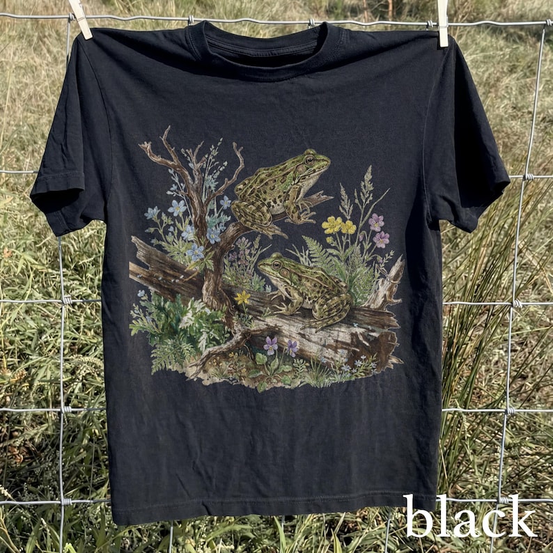 May include: Black t-shirt featuring a detailed illustration of two frogs perched on a log surrounded by plants and flowers. The design includes a variety of colors, including green, blue, and yellow. The word "black" is printed at the bottom of the shirt.