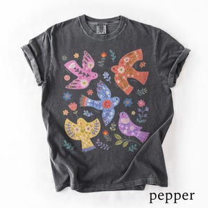 May include: A charcoal grey short-sleeve t-shirt featuring a graphic print of four stylised birds in pink, orange, blue, and purple, surrounded by small floral and leaf motifs. The shirt has a crew neckline and a washed, vintage-style finish.