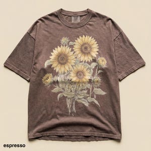Pode incluir: Camiseta cor de café com uma ilustração de girassóis. O design apresenta vários girassóis em plena floração, botões e folhas. A camisa tem um corte folgado e mangas curtas. A palavra "espresso" está impressa na parte inferior.