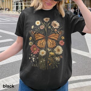 Puede incluir: Camiseta negra con una ilustraci&oacute;n detallada de una mariposa monarca con alas naranjas y negras, rodeada de flores coloridas. El texto "black" est&aacute; impreso en la parte inferior.