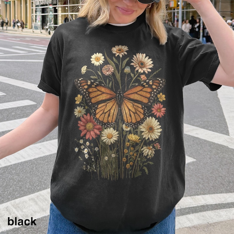 Puede incluir: Camiseta negra con una ilustraci&oacute;n detallada de una mariposa monarca con alas naranjas y negras, rodeada de flores coloridas. El texto "black" est&aacute; impreso en la parte inferior.