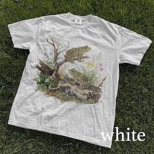 May include: White t-shirt featuring a detailed illustration of two green frogs perched on a log with surrounding plants and flowers. The shirt has a classic crew neck and short sleeves. The word "white" is printed at the bottom of the shirt.