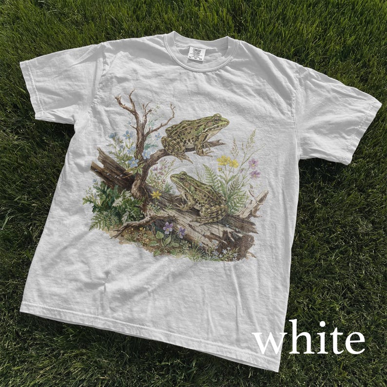 May include: White t-shirt featuring a detailed illustration of two green frogs perched on a log with surrounding plants and flowers. The shirt has a classic crew neck and short sleeves. The word "white" is printed at the bottom of the shirt.
