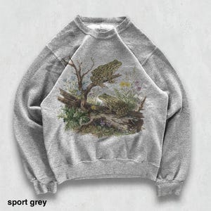 May include: Sport grey sweatshirt featuring a detailed illustration of two frogs perched on a log with foliage. The graphic includes a tree branch, flowers, and the text "sport grey" at the bottom. The sweatshirt has a crew neck and long sleeves.