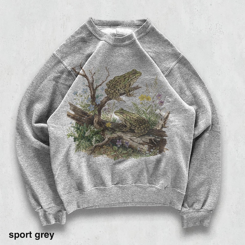 May include: Sport grey sweatshirt featuring a detailed illustration of two frogs perched on a log with foliage. The graphic includes a tree branch, flowers, and the text "sport grey" at the bottom. The sweatshirt has a crew neck and long sleeves.