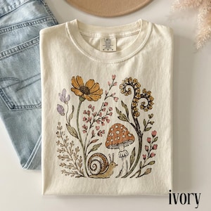 May include: An ivory t-shirt featuring a graphic print of a brown snail, a spotted mushroom, and various wildflowers in shades of yellow, pink, and green. The inner neck label displays the text Comfort Colors and size M-M-M.