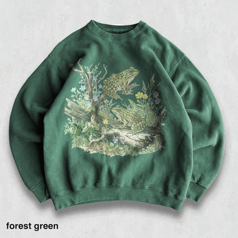 May include: Forest green sweatshirt featuring a detailed graphic of two frogs perched on a log amidst foliage and flowers. The design includes a tree branch and various plants. The text "forest green" is at the bottom.