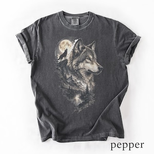 Könnte beinhalten: Ein anthrazitfarbenes Kurzarm-T-Shirt mit einem detaillierten Grafikdruck eines Wolfskopfes, eines kleineren heulenden Wolfes vor einem Vollmond und einer Bergwaldlandschaft. Das Shirt hat einen Rundhalsausschnitt und eine verwaschene Optik.