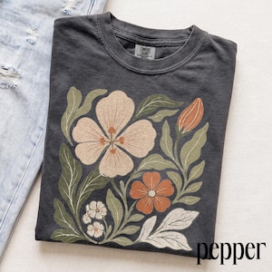 May include: A charcoal gray crew neck t-shirt featuring a graphic print of various flowers and leaves in muted tones of cream, terracotta, and sage green. The shirt has a Comfort Colors brand label at the inner neckline and is folded next to light blue jeans.