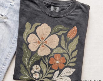 May include: A charcoal gray crew neck t-shirt featuring a graphic print of various flowers and leaves in muted tones of cream, terracotta, and sage green. The shirt has a Comfort Colors brand label at the inner neckline and is folded next to light blue jeans.