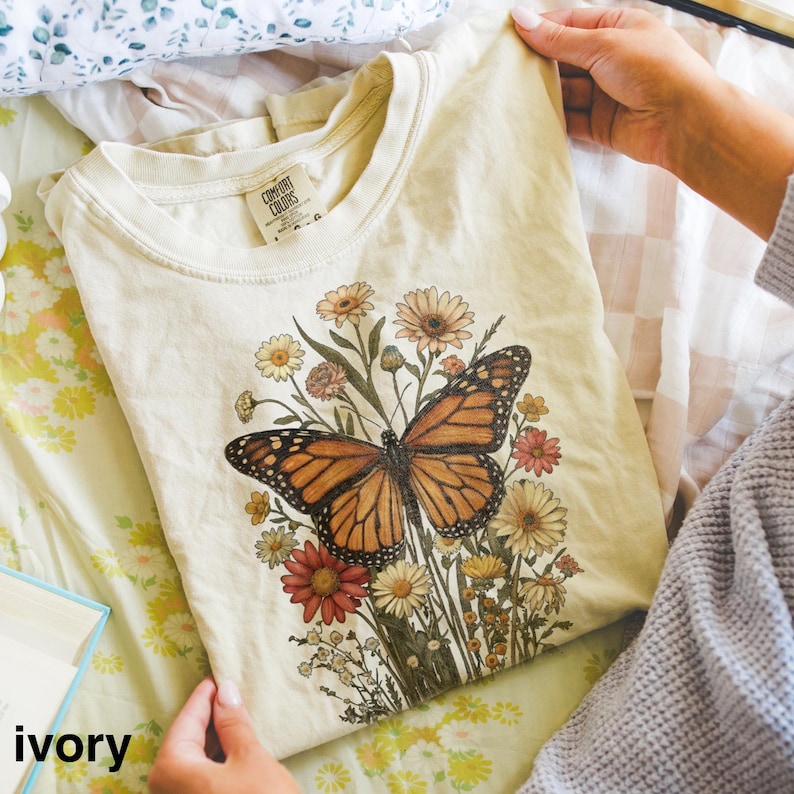Puede incluir: Camiseta de color marfil con una mariposa monarca con alas naranjas y negras, rodeada de flores coloridas. La camiseta est&aacute; hecha de un material suave y es de la marca Comfort Colors.