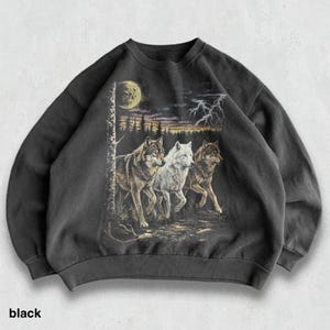 May include: Black sweatshirt featuring a graphic of three wolves running through a forest under a moonlit sky. The design includes a tree, lightning, and a color palette of black, brown, white, and yellow.