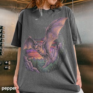 May include: Charcoal grey t-shirt with a bat graphic in shades of purple and brown, set against a sunset. The bat is flying over rocky terrain. The word "pepper" is visible at the bottom of the image.