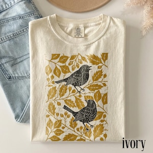 May include: An ivory t-shirt featuring a graphic print of two black birds perched among mustard yellow leaves and branches. The shirt has a crew neckline and a Comfort Colors brand label on the inside collar. The fabric has a soft, textured appearance.