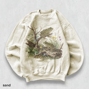 May include: Cream-coloured sweatshirt with a detailed illustration of two frogs on a log surrounded by plants and wildflowers. The design includes a tree branch, creating a nature-themed image. The word "sand" is printed at the bottom of the sweatshirt.