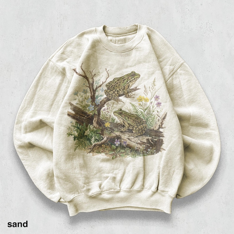 May include: Sand-colored sweatshirt featuring a detailed illustration of two frogs perched on a log amidst foliage and wildflowers. The artwork includes a tree branch and various plants, creating a nature-themed design. The word "sand" is printed at the bottom.