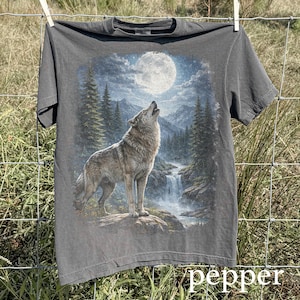 May include: A charcoal gray t-shirt featuring a detailed graphic of a howling wolf under a full moon. The design includes a waterfall, pine trees, and a mountain range. The shirt is hanging on a clothesline.