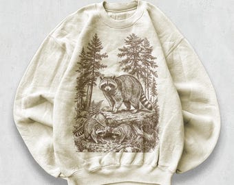 May include: Sand-colored sweatshirt featuring a detailed illustration of raccoons in a forest setting. The design includes three raccoons, trees, and foliage, creating a nature-themed graphic. The word "sand" is printed at the bottom.
