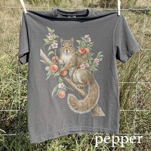 May include: A charcoal gray t-shirt featuring a detailed illustration of a squirrel perched on a tree branch with peaches and pink blossoms. The word "pepper" is printed in white at the bottom of the shirt. The shirt is hanging on a clothesline.