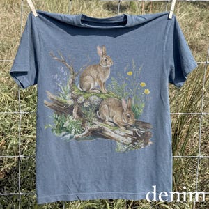 K&ouml;nnte beinhalten: Denimblaues T-Shirt mit einer detaillierten Illustration von zwei Kaninchen inmitten von Wildblumen und Laub. Das Kunstwerk ist auf der Vorderseite aufgedruckt. Das Wort "denim" ist unten auf dem T-Shirt in Wei&szlig; aufgedruckt.