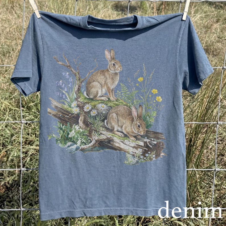 K&ouml;nnte beinhalten: Denimblaues T-Shirt mit einer detaillierten Illustration von zwei Kaninchen inmitten von Wildblumen und Laub. Das Kunstwerk ist auf der Vorderseite aufgedruckt. Das Wort "denim" ist unten auf dem T-Shirt in Wei&szlig; aufgedruckt.